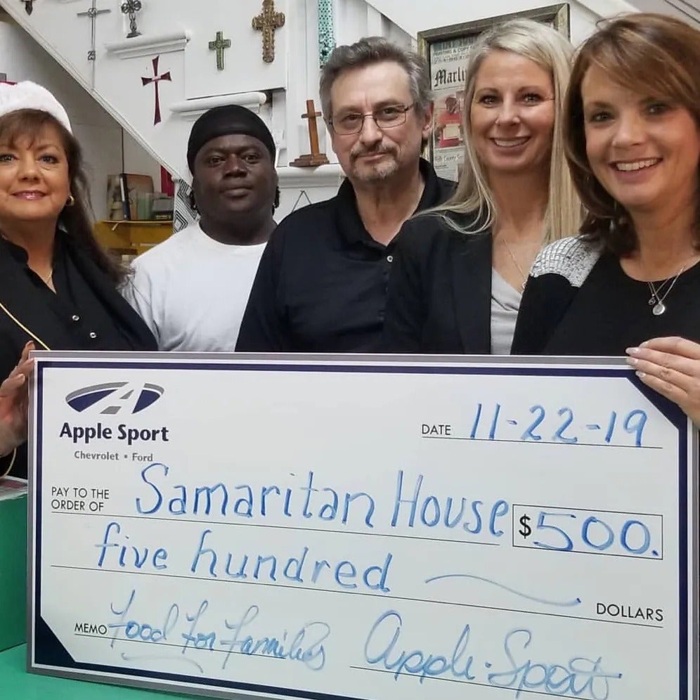 A group of people holds a large check from Apple Sport Chevrolet Ford donating $500 to Samaritan House for food for families.